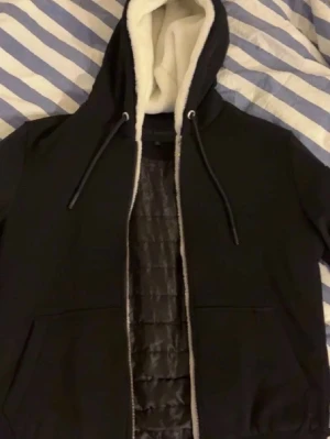 Svart hoodie med foder och dragkedja - Mysig svart hoodie med vit fluffig fleecefoder i huvan och dragkedja framtill. Utrustad med två stora fickor och justerbara snören i huvan. Insidan har quiltat foder för extra värme. Perfekt för kyliga dagar.