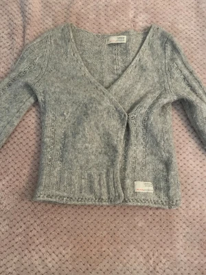 Grå stickad kofta från Odd Molly - Mysig grå stickad kofta från Odd Molly med v-ringning och ribbade detaljer. Koftan har en figurnära passform och en liten etikett med logga nedtill.