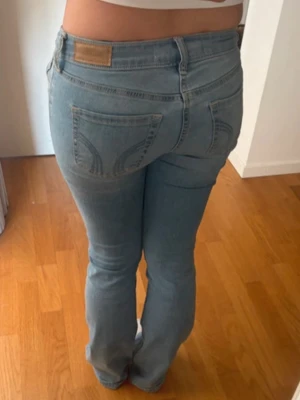 Hollister jeans - Säljer ett par ljusblå jeans med slitningar på låren och knäna. Modellen är bootcut med normalhög midja och klassiska fem fickor. Jeansen har snygga sömmar på bakfickorna och stängs med knapp och dragkedja. Perfekta för en trendig och avslappnad look.