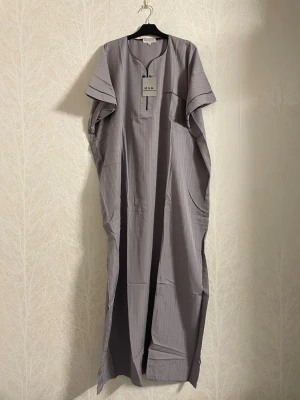 Grå Thobe från Saudiarabien - Thobe i jättefin silvergrå färg i storlek 56, köpt från Saudiarabien. Har aldrig använts och sitter fortfarande i sin originala förpackning. Har fickor  vid sidorna, en bröstficka, och knäppning framtill. Materialet är mjukt och svalt, och passar såväl till hemmet som till utomhus!   Längd: ca 137 cm.  Axel: ca 37 cm.