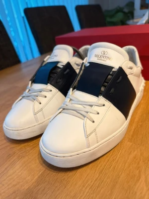 Valentino vita sneakers med blå detalj - Snygga vita sneakers från Valentino med bred mörkblå läderdetalj över mitten och klassiska vita snören. Skorna har en låg profil och stilren design med logga på plösen. Perfekta för dig som vill ha en exklusiv touch till din outfit.