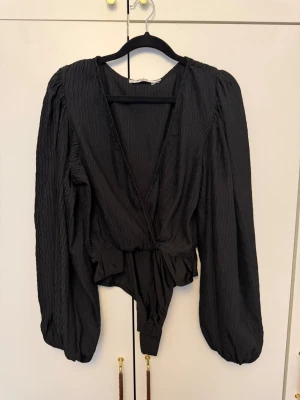 Svart body med puffärm från NA-KD - Säljer en svart body från NA-KD med djup v-ringning och långa puffärmar. Materialet är lätt glansigt och har en strukturerad yta. Modellen har omlott-design och markerad midja som ger en snygg siluett. Perfekt för dig som vill ha något trendigt och stilrent.