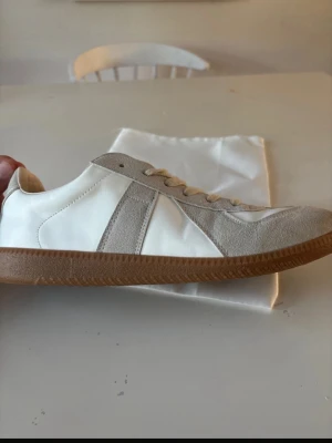 Vita och beige Maison margielas gats - Snygga sneakers i vitt och beige med ovandel i mocka och slät textil från Maison margiela. Skorna har klassisk låg profil, vita snören och en brun gummisula som ger en retrovibe. Perfekta för dig som gillar stilrena och tidlösa sneakers.