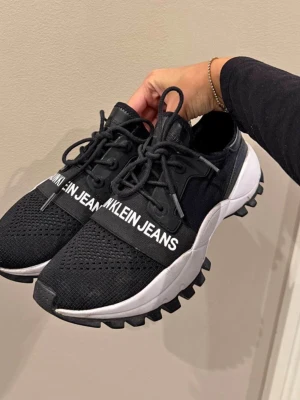 Svarta sneakers Calvin Klein Jeans - Snygga svarta sneakers från Calvin Klein Jeans med vit sula och grovt mönstrad yttersula. Skorna har snörning och ett brett band över foten med loggan tryckt i vitt. Ovandelen är i mesh och syntetmaterial för en sportig look.
