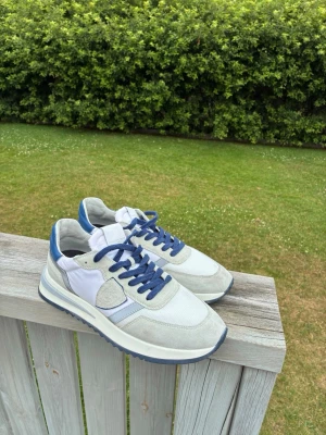 Vita och blå Philippe Model sneakers - Snygga sneakers från Philippe Model i vitt och blått med detaljer i mocka och mesh. Skorna har blå snören, vit sula med blå kant och en cool logga på sidan. Perfekta för dig som gillar stilrena och sportiga skor med en modern touch. Säljer nästan helt nya Philippe model skor som bara suttit hemma utan användning. Köpte helt nya på nätet. Kan lösa kvitto vid begäran.