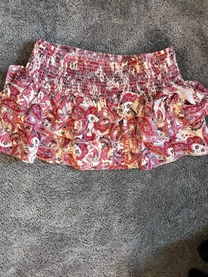 Rosa och röd volangkjol med paisleymönster - Söt kortkjol med volanger och paisleymönster i rosa och rött. Kjolen har smock upptill för en bekväm passform och är perfekt för dig som gillar färgglada plagg med mycket detaljer. Materialet känns lätt och luftigt.