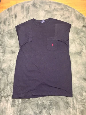 Svart t-shirt från Polo Ralph Lauren - Svart t-shirt från Polo Ralph Lauren med klassisk passform och korta ärmar. T-shirten har en bröstficka med det ikoniska röda Polo-logot broderat på. Materialet är mjuk bomull och perfekt för en avslappnad look.