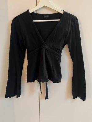 Svart omlottopp med knyt från GinaTricot  - Svart omlottopp från GinaTricot med v-ringning och långa ärmar. Toppen har knytband i midjan som kan knytas baktill för en snygg passform. Materialet är lätt och har en diskret struktur. Perfekt för att skapa en cool och stilren look.