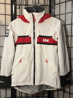 Helly Hansen Salt Inshore Sailing Jacket – Helly Tech Protection – M -   Helly Hansen Salt Inshore Sailing Jacket (Model 30283) i mycket fint skick.  Teknisk seglarjacka med Helly Tech Protection-membran som gör den vatten- och vindtät samtidigt som den andas. Klassisk HH sailing-design i vit, röd och svart färgkombination.  Jackan är från Helly Hansens populära Salt-serie, framtagen för kust- och fritidssegling. Fungerar lika bra för outdoor, regn eller som clean streetwear.  Skick   Nära nyskick.