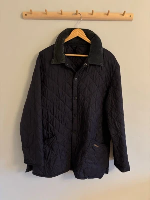 Svart quiltad jacka från Barbour - Klassisk svart quiltad jacka från Barbour med tryckknappar framtill och två stora fickor. Jackan har en bred krage i manchestertyg och ett tidlöst rutmönster i quiltningen. Perfekt för dig som gillar stilren och funktionell design.