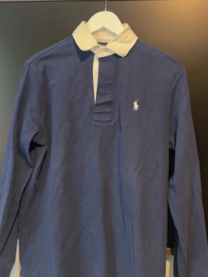 Marinblå långärmad polotopp Polo Ralph Lauren - Klassisk marinblå långärmad polotopp från Polo Ralph Lauren med vit krage och broderad logga på bröstet. Tillverkad i mjuk bomull och har knappar vid halsen. Perfekt för en stilren och sportig look. Svår att få tag på då denna variant är slutsåld!