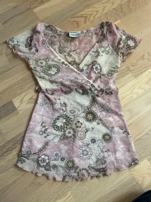 Blommig omlottopp från Clockhouse - Supersöt omlottopp i tunn mesh från Clockhouse med blommigt mönster i rosa, beige och grönt. Korta ärmar, volangkant och knytband i midjan. Superfin på sommaren med en kjol eller ett par snygga jeans! 