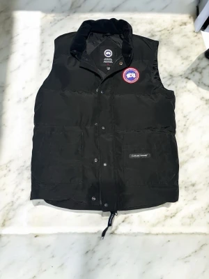 Svart dunväst från Canada Goose - Snygg svart dunväst från Canada Goose med klassisk logotyp på bröstet och två stora fickor framtill. Västen har hög krage, tryckknappar och dragsko nedtill. Perfekt för lager-på-lager och håller dig varm under kyliga dagar.