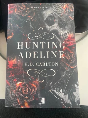 Hunting Adeline av H.D. Carlton - En mörk och spännande skönlitterär bok med titeln 'Hunting Adeline' av H.D. Carlton. Omslaget är svart med röda rosor, spindelnät, en dödskalle och en fjäril, vilket ger en mystisk och dramatisk känsla. Perfekt för dig som gillar thrillers och romantik. Och den är på polska 