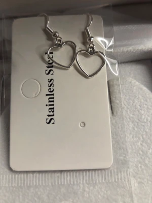 Silverfärgade hjärtörhängen i stål - Säljer ett par stilrena örhängen i form av öppna hjärtan. De är silverfärgade och tillverkade i rostfritt stål, vilket gör dem både snygga och tåliga. Perfekta för dig som gillar minimalistisk design och vill ha något enkelt men ändå gulligt.
