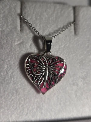 Halsband med hjärthänge och fjäril som lyser i mörker  - Unikt halsband med ett hjärtformat hänge i silverfärgad metall. Hänget har ett detaljerat fjärilsmotiv och byter färg från rosa i dagsljus till lila i mörker. Vanligt dagsljus aktiverar effekten i hänget och det börjar därför att lysa/ge ett lila sken i mörker som kan vara i flera timmar. Perfekt statement-piece för dig som gillar smycken med coola effekter och snygg design. Kedjan är 45 cm + 5 cm förlängningskedja i rostfritt stål. Handgjort halsband. 