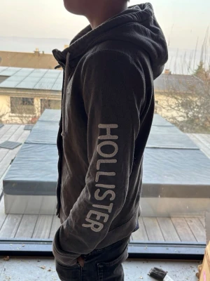 Mörkgrå hoodie från Hollister - Snygg mörkgrå hoodie från Hollister med dragkedja och knappar framtill. Stor logga på ärmen och liten logga på bröstet. Utrustad med huva och vita dragsnören samt två fickor framtill. Perfekt för en avslappnad och trendig stil.