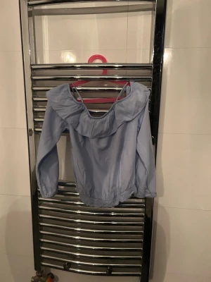 Ljusblå offshoulder blus - Trendig ljusblå offshoulder blus med bred volang runt axlarna och långa ärmar. Blusen har resår i nederkant och vid ärmsluten för en snygg passform. Perfekt att styla med jeans eller kjol för en fräsch look.