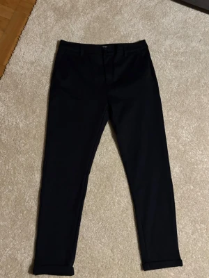 Svarta kostymbyxor från SMOG - Stilrena svarta kostymbyxor från SMOG med smal passform och uppvikta benslut. Klassisk design med fickor i sidan och hällor för bälte. Perfekta för dig som vill ha en clean och modern look.