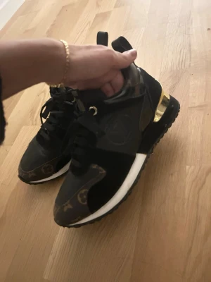 Svarta Louis Vuitton sneakers med guld - Snygga svarta sneakers från Louis Vuitton med klassiskt monogrammönster på tå och häl, samt gulddetaljer på hälen. Skorna har svarta skosnören, vita sulor och är tillverkade i en mix av läder och textil. Perfekt för dig som vill ha något exklusivt och trendigt. Dom är lite förstora för mig då jag har storlek 37. 