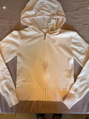 Ljusrosa hoodie från Gina Tricot - Helt ny. Mjuk och skön ljusrosa hoodie från Gina Tricot med dragkedja framtill och huva. Tillverkad i stickad bomullsblandning som känns riktigt bekväm. Perfekt för dig som gillar en enkel och clean stil. Långa ärmar och ribbade muddar för extra komfort.