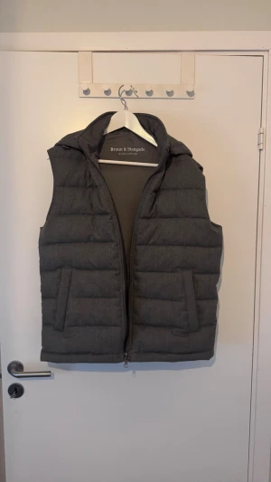 Mörkgrå väst med huva - Snygg mörkgrå dunväst från Brun & Stengade med quiltad design och praktisk huva. Västen har två stora fickor framtill och dragkedja hela vägen. Perfekt för lager-på-lager och ger en clean look till din outfit. Nypris 2199kr