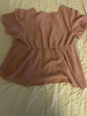 Rosa blus med prickigt mönster - Söt rosa blus med små vita prickar, v-ringning och knappar framtill. Blusen har korta ärmar med lätt volang och markerad midja för en feminin siluett. Perfekt till jeans eller kjol för en trendig look.