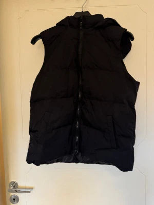 Svart dunväst med huva - Säljer en svart dunväst med huva och dragkedja framtill. Västen har två fickor på sidorna och en enkel, stilren design. Perfekt för lager-på-lager och funkar till många olika outfits. Storlek S/M. 