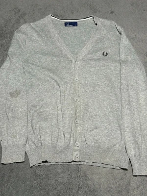 Fred Perry Cardigan Grey – Laurel Wreath Logo – Size S - Klassisk Fred Perry cardigan i grått med broderad laurel wreath-logo på bröstet.  Tunn och mjuk stickning som passar perfekt för lager på lager året runt. Tidlös brittisk stil.  Detaljer • Fred Perry • Grå cardigan • Broderad laurel logo • Knäppning framtill • Storlek S • 100% bomull  Skick: bra begagnat skick. En mindre fläck på ena ärmen (se bild).