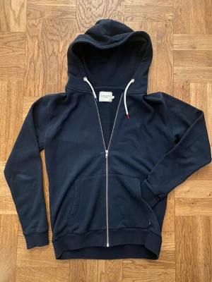 Marinblå zip-hoodie från Les Deux - Snygg marinblå hoodie från Les Deux med dragkedja framtill, stor huva och vita dragsnören. Hoodien har två rymliga fickor fram och ribbade muddar vid ärmar och nederkant. Perfekt för en avslappnad och stilren look.