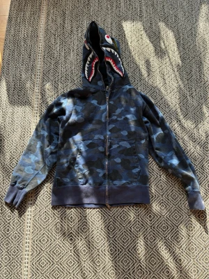 Blå camo hoodie från BAPE - Säljer en blå camo zip hoodie från BAPE med ikonisk hajdesign på huvan och broderade detaljer i orange, rött och vitt. Hoodien har fickor fram, ribbade muddar och dragkedja. Storlek M men passar som en S/XS.