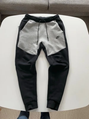 Nike Tech Fleece mjukisbyxor svart/grå - Snygga Nike Tech Fleece mjukisbyxor i svart och ljusgrått med dragsko i midjan och ribbade muddar vid bensluten. Köpta från nike.com för 1259 kr och är i nästan helt nytt skick, inga fläcker eller andra defekter. Säljer dem pga att jag inte använder dem. Byxorna har två stora fickor fram och en dragkedjeficka bak. Perfekta för chill eller träning, tillverkade i mjukt och stretchigt material.