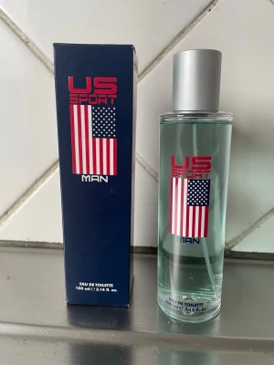 US Sport Man Eau de Toilette 100 ml - US Sport Man Eau de Toilette är en fräsch herrparfym i en stilren, genomskinlig glasflaska med silverfärgat lock. Flaskan och kartongen har en amerikansk flagga och röd text. Innehåller 100 ml.