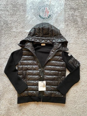 Moncler cardigan  - Snygg svart dunväst från Moncler med stickade svarta ärmar och stor huva. Västen har två fickor med dragkedja och ikonisk Moncler-logga på ärmen. Materialet är glansigt dun på kroppen och mjukt stickat tyg på ärmarna. Perfekt för lager-på-lager och streetstyle.