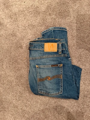 Blå jeans från Nudie Jeans - Säljer ett par klassiska blå jeans från Nudie Jeans med snygga slitningar och kontrastsömmar. Jeansen har fem fickor, en cool broderad detalj på bakfickan och en läderpatch med logotyp. Perfekt för dig som gillar en avslappnad och trendig look.