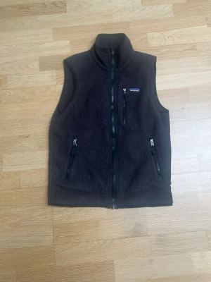 Svart fleeceväst från Patagonia - Mysig svart fleeceväst från Patagonia med hög krage och dragkedja framtill. Västen har tre fickor med dragkedja och en klassisk, enkel design. Perfekt för lager-på-lager och chill stil.