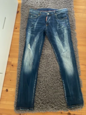 Blå slim fit jeans med slitningar - Säljer ett par blå slim fit jeans med slitningar och ljusa tvättade partier på benen. Jeansen har fem fickor, knappgylf och detaljer med färgstänk för en trendig look. Perfekta för dig som gillar en avslappnad och modern stil.