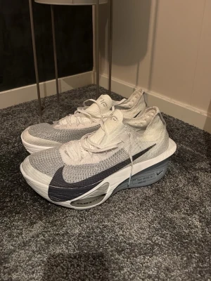 Nike Alphafly 3 vit/grå - Säljer ett par Nike löparskor efter endast EN löptur!. Skorna är i vitt och grått med stor svart swoosh på sidan. Skorna har ventilerande mesh-ovandel och avancerad Flyplate-teknologi i sulan för extra energiåtergivning. Snygg, sportig design och låg profil som passar perfekt till träning eller streetwear.