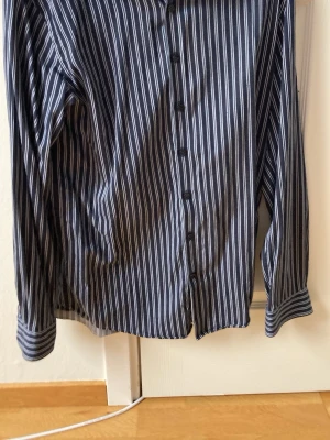 Randig skjorta från Lindbergh - Snygg mörkblå skjorta från Lindbergh med vita vertikala ränder. Klassisk krage, långa ärmar och knäppning framtill. Perfekt för dig som gillar stilrena och tidlösa plagg.