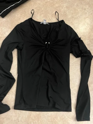 Svart långärmad topp med metall - Trendig svart långärmad topp med smala axelband och snygg snörningsdetalj framtill som ger en cool twist. Toppen har en figurnära passform och är perfekt att styla med både jeans och kjol. Materialet känns mjukt och stretchigt.
