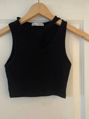 Svart ribbad crop top Pull&Bear - Säljer en svart ribbad crop top från Pull&Bear med asymmetrisk cut-out vid axeln. Toppen är ärmlös och har en tight passform som framhäver figuren. Perfekt till jeans eller kjol för en trendig look.