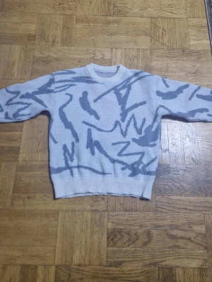 Ljusgrå stickad tröja med mönster - Säljer en ljusgrå stickad tröja med abstrakt mönster i mörkare grå toner. Tröjan har rund halsringning, långa ärmar och ribbstickade muddar vid ärmslut och nederkant. Perfekt för dig som gillar en unik och trendig look.