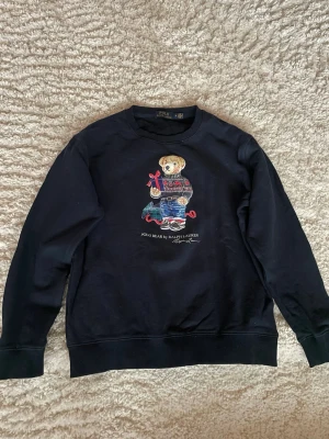 Mörkblå sweatshirt Polo Bear Ralph Lauren - Svart sweatshirt från Ralph Lauren med ikoniska Polo Bear-trycket framtill. Björnen har på sig en julig stickad tröja, jeans och håller i en present. Rund halsringning och ribbade muddar. Perfekt för dig som gillar klassiska streetwear-vibbar. Sitter mer som en S.