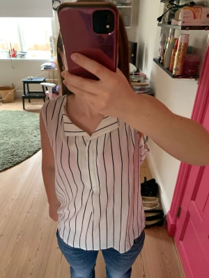 Vit randig ärmlös linnetopp - Säljer en vit ärmlös linnetopp med svarta vertikala ränder och snygg krage. Toppen har en avslappnad passform och knäppning framtill. Perfekt till jeans eller kjol för en fräsch look.