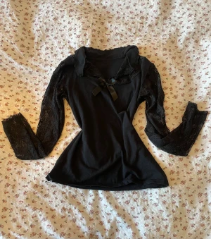 Svart topp med spetsärmar och rosett - Supersnygg svart topp med långa spetsärmar och en söt rosett framtill vid halsringningen. Toppen har en feminin passform och spetsdetaljer både vid ärmar och urringning. Perfekt för dig som gillar en lite romantisk och edgy stil~ Okänt märke och ingen storlek tyvärr men skulle säga att den passar xs! 🤗🖤