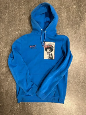 Supply & Demand - Cool blå hoodie från Supply and Demand med stor huva och dragsko. På bröstet finns ett svartvitt tryck av Frihetsgudinnan och röda detaljer samt text. Hoodien har en klassisk känguruficka och ribbade muddar vid ärmar och nederkant. Perfekt för dig som gillar streetwear.