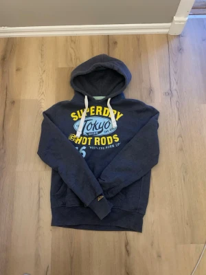 Mörkblå Superdry hoodie Tokyo print - Snygg mörkblå hoodie från Superdry med stort Tokyo Hot Rods-tryck i gult, blått och vitt på bröstet. Klassisk huva med vita snören och mjukt, borstat innerfoder. Limited Edition Vintage Goods, perfekt för dig som gillar streetwear och unika plagg.