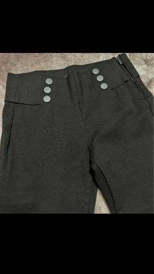 Svarta leggings med knappar - Snygga svarta leggings med hög midja och dekorativa mattsvarta knappar framtill. Byxorna har en smal passform och dragkedja i sidan för en clean look. Materialet ser ut att vara stretchigt och bekvämt, perfekt för en stilren outfit.   Bredd - ca 37cm Innelängd - ca 64cm Längd - ca 88cm
