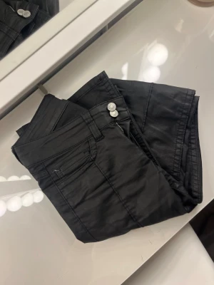 Svarta coated jeans med bootcut - Säljer ett par svarta coated jeans med bootcut-modell och dubbla silverknappar i midjan. Jeansen har klassisk femficksdesign och en glansig finish som ger en cool look. Perfekta för dig som vill ha ett par trendiga och snygga jeans.