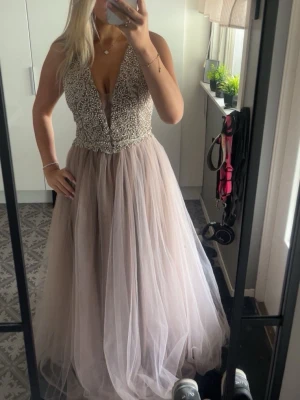 Lång champagnefärgad balklänning - Säljer en elegant långklänning i champagnefärg med djup v-ringning och pärlbroderad överdel. Kjolen är i flera lager tyll som ger en fluffig och drömmig look. Perfekt för bal eller festliga tillfällen.